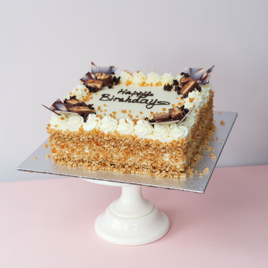 Square birthday cake with white frosting, chocolate decorations, and 'Happy Birthday' text on a pink surface.