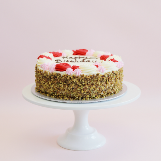 Birthday cake with white frosting, red flowers, and pink roses on a white cake stand against a light pink background.
